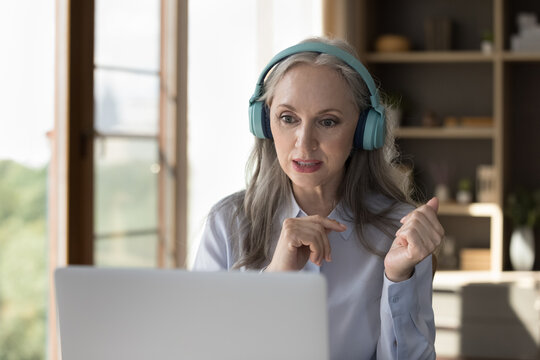 Focused engaged senior laptop user woman in wireless big headphones making video call, watching webinar, media content, studying at home. Online teacher, blogger presenting training, coaching - Powered by Adobe