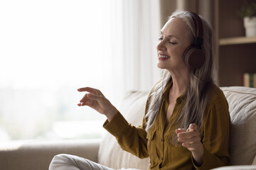 Happy joyful beautiful elderly lady in modern headphones having fun at home, listening to hifi...
