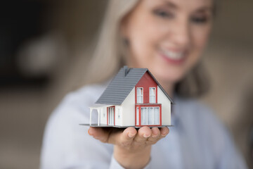 Happy realtor woman holding tiny house model on hand, palm, showing object at camera, promoting agency service, help with house, property buying apartment rent, mortgage