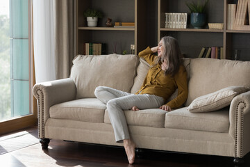 Obraz premium Happy beautiful grey haired elderly woman sitting on comfortable couch in living room, leaning on back, relaxing, thinking, dreaming with happy smile, enjoying leisure, being at home