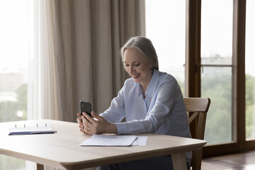 Happy elderly retired smartphone user talking on video call, enjoying online conversation, holding mobile phone over table with papers at home, smiling, laughing. Retirement, communication concept