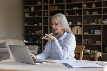 Elderly retired business woman working from home, using laptop, recording voice message for online...
