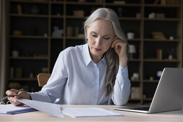 Focused busy elderly freelance business woman doing paperwork, accounting job, reading documents at laptop, checking insurance, loan, mortgage, rent agreement, reviewing bills