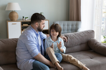 Loving dad listen with interest his little son, sit on sofa spend time together, enjoy conversation, trustworthy communication having warm friendly relations. Understanding, family, fatherhood concept