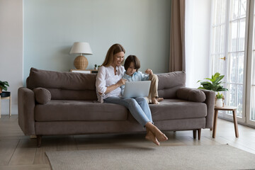 Young mom and cute little 6s son sit on sofa with laptop spend leisure watch videos, buy goods on-line. Child education, development using modern tech and internet, e-commerce shopping at home concept