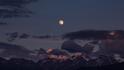 A full moon rising at dusk, just after sunset across a snow capped mountain range