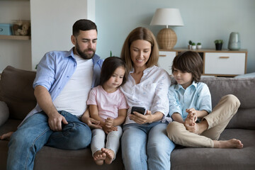 Young couple and preschoolers pretty children enjoy new mobile app using smartphone, make call or buying goods on-line, spend pastime on internet together resting on sofa at home. Modern tech concept