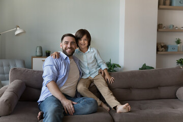 Loving preschooler son hugging young father sit on sofa smile feel happy enjoy leisure together at modern home, pose shooting for camera. Two generation people, good friendly relations, love concept