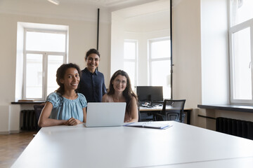 Young multiracial women colleagues working on new collaborative project use computer smile look at camera, meet in coworking. Friendly relation, motivation, effective workflow use modern tech concept