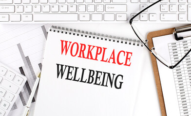 Text Workplace Wellbeing on Office desk table with keyboard, notepad and analysis chart on white...