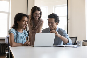 Three multiracial colleagues use laptop speaking on project, discuss collaborative task, learn new software, enjoy amusing content during break at workplace. Teamwork, cooperation, modern tech concept