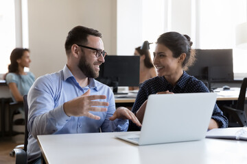 Millennial Indian woman and Caucasian man colleagues smile while working on project sit in co-working workspace with laptop. Teamwork, friendly relations at workplace, tech, creative workflow concept