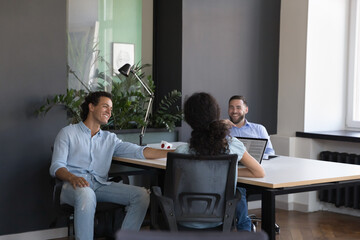 Three diverse office employees joking talking during break at workplace sit at shared desk looking happy have fun share creative ideas working together in co-working. Good friendly relations concept