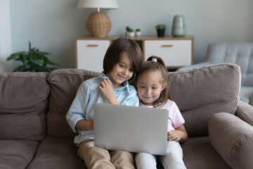 Preschooler children sit on sofa with laptop spend time on internet, enjoy favourite video vlog,...