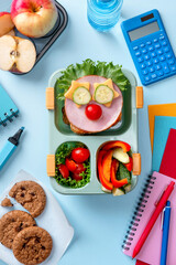 School lunch box for kids with food in the form of funny faces. School lunch box with sandwich, vegetables, water and stationery on table. Healthy eating habits concept. Back to school concept