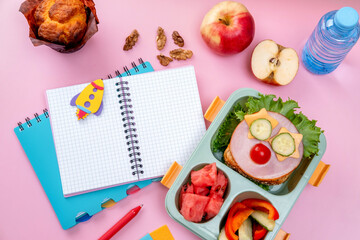 School lunch box for kids with food in the form of funny faces. School lunch box with sandwich, vegetables, water and stationery on table. Healthy eating habits concept. Back to school concept
