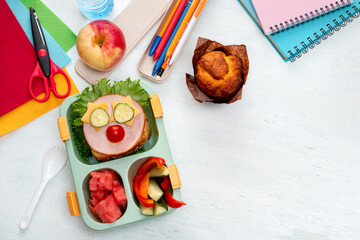 School lunch box for kids with food in the form of funny faces. School lunch box with sandwich, vegetables, water and stationery on table. Healthy eating habits concept. Back to school concept