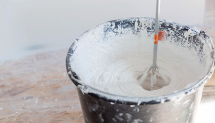 A repair worker mixes putty with water in a bucket using a hand mixer for building mixes.