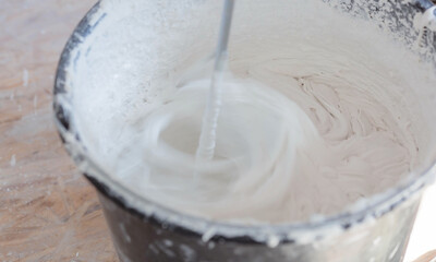 A repair worker mixes putty with water in a bucket using a hand mixer for building mixes.