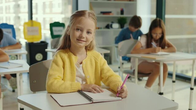 Portrait of a beautiful blonde pupil schoolgirl sitting on a chair in the classroom against the background of children holding a pen to write something down. People and educational concept.