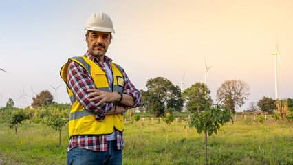 Naklejka na ściany i meble Aืn Engineer standing by the wind turbine in the renewable power farm. He is wearing a white hard hat and yellow transparent vest.