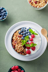 Greek yogurt with raspberries, blueberries and granola on a green background. Bowl with healthy breakfast on white plate with wooden spoon. Top view. Copy space