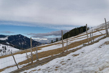 A snow-covered steep alpine meadow with a wooden fence. The deep blue sky is partially covered and...