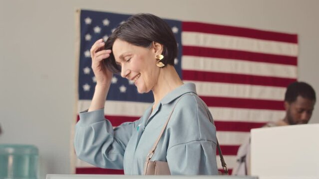 Medium slowmo of friendly mature Caucasian woman talking to people working at voting station while taking her bulletin and going to vote booth