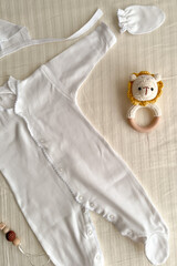 a white children's suit and a rattle with a lion are lying on a light bed. View from above. Clothes...
