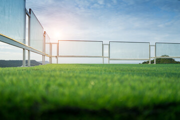 Focus at glass fence around the green grass field with sunlight and blue sky background at the terrace.