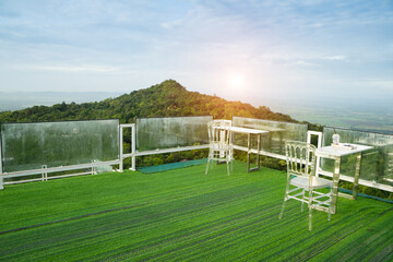 Outdoor living terrace with artificial green grass ,chair and table around with glass guard rail  with mountain and sunlight background.