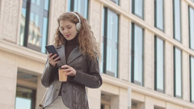 Low angle medium slowmo of attractive young smiling woman in headphones scrolling on smartphone standing outdoors against tall beige building with takeaway coffee cup in hand