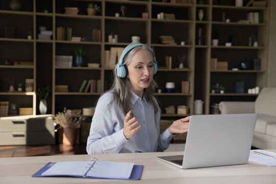 Busy Middle Aged Online Teacher In Wireless Headphones Speaking At Laptop Webcam, Holding Online Conference, Making Video Call At Home Office Workplace. Communication Concept