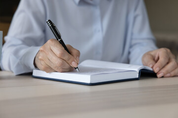 Middle aged business woman wearing pale blue shirt, writing notes in notebook at work table. Hands...