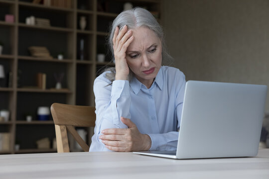 Upset unhappy mature business lady thinking over bad news at laptop, sitting at workplace table with computer, holding head, suffering from headache attack, feeling tired, frustrated