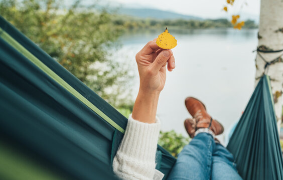 Young woman holding in fingers and examining small yellow birch tree leaf as she swinging in hammock on the mountain lake bank. Out-of-town Outdoor Recreation in Nature concept image.