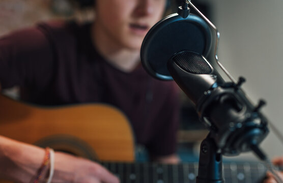 Microphone with pop filter closeup photo with a young teenager boy in headphones recording voice and guitar playing music. Modern home sound studio audio recording technology and teens hobby concept.