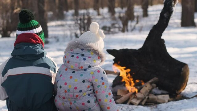 Two children in the winter forest sit by the fire back view. Children bask near the fire in snowy forest.
