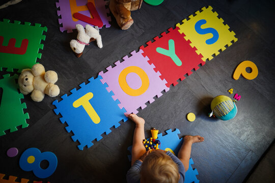 bird view of colorful kids puzzle mat playground in nursery or at home with the letters TOYS on it lying on the floor while one year old blond baby in white body is playing