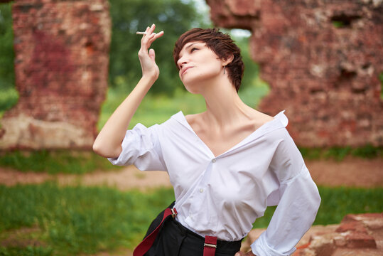 Image of a stylish beautiful woman in a shirt, trousers and suspenders who smokes a cigarette in the park against the backdrop of a destroyed building. The concept of style and fashion.
