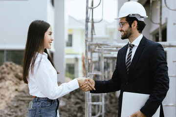A young white engineer and an Asian woman designing a house hold hands at a construction site.