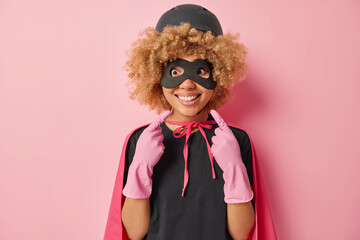 Photo of cheerful curly haired young woman points at toothy smile shows white teeth wears helmet eyemask rubber gloves cloak has glad expression comes on masquerade isolated over pink background.