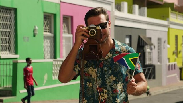 tourist man smiling happy using vintage camera at the city. man taking photographs on SLR. stylish man in a summer shirt makes a photo against the background of colored houses and smiles at the camera