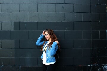 Portrait of a woman touching her hair. Beautiful young girl wearing a jean jacket and a white t-shirt. Girl with black glasses and earings. Black wall background. Long, shiny and curly ginger hair. - Powered by Adobe