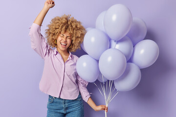 Young cheerful woman dressed in shirt and jeans holds bunch of inflated helium balloons celebrates...