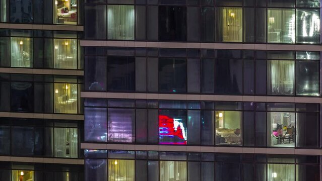 Apartment windows with blinds of a glazed skyscraper glow at night with city lights reflection aerial timelapse. Illuminated towers