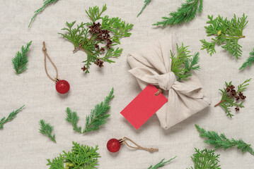 A Christmas gift wrapped in cloth with a blank label and Christmas decorations. Beige linen textile background. Gift in furoshiki style. Zero waste concept. Top view, selective focus.