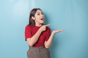 Shocked Asian woman wearing red t-shirt pointing at the copy space beside her, isolated by blue background