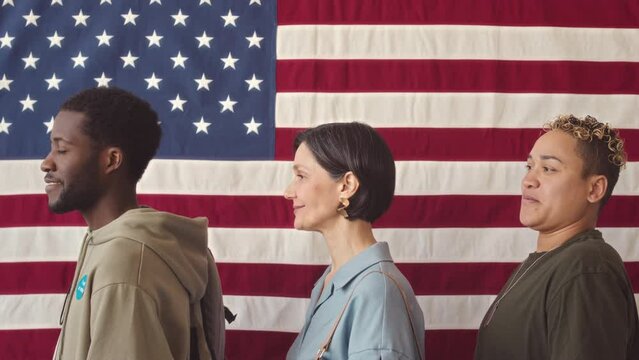 Waist up slowmo portrait of diverse US citizens standing in queue against US flag on wall waiting for voting procedure