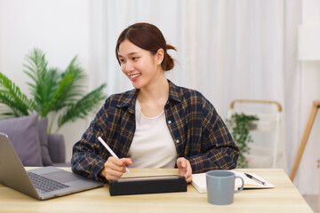 Lifestyle in living room concept, Young Asian woman using laptop and taking notes data on tablet